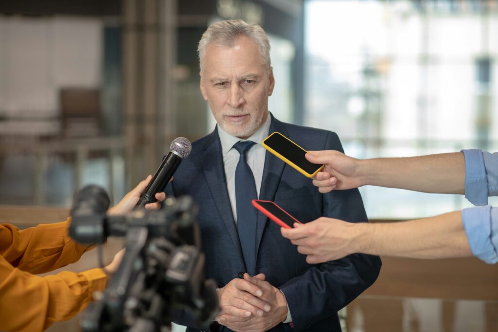 A grey-haired businessman in a navy suit stands with his hands clasped and a wary expression, facing a microphone, a TV camera and two smartphones held out by reporters.